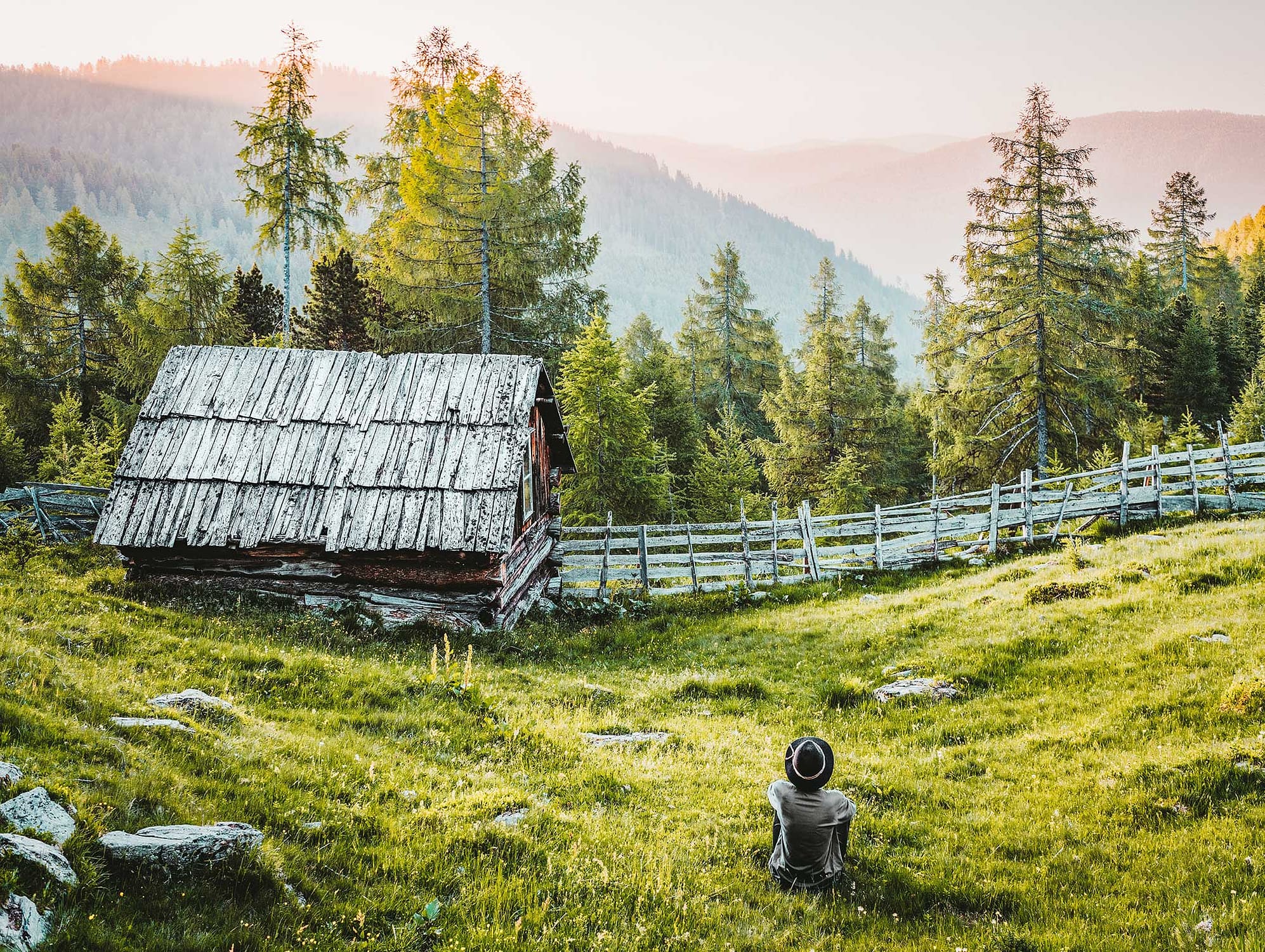 a man sits in a field with an old building and fence overlooking some mountains and trees