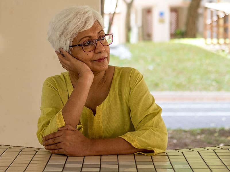 A middle-aged woman sits at a table with her head resting on her hand looking pensively away from the camera