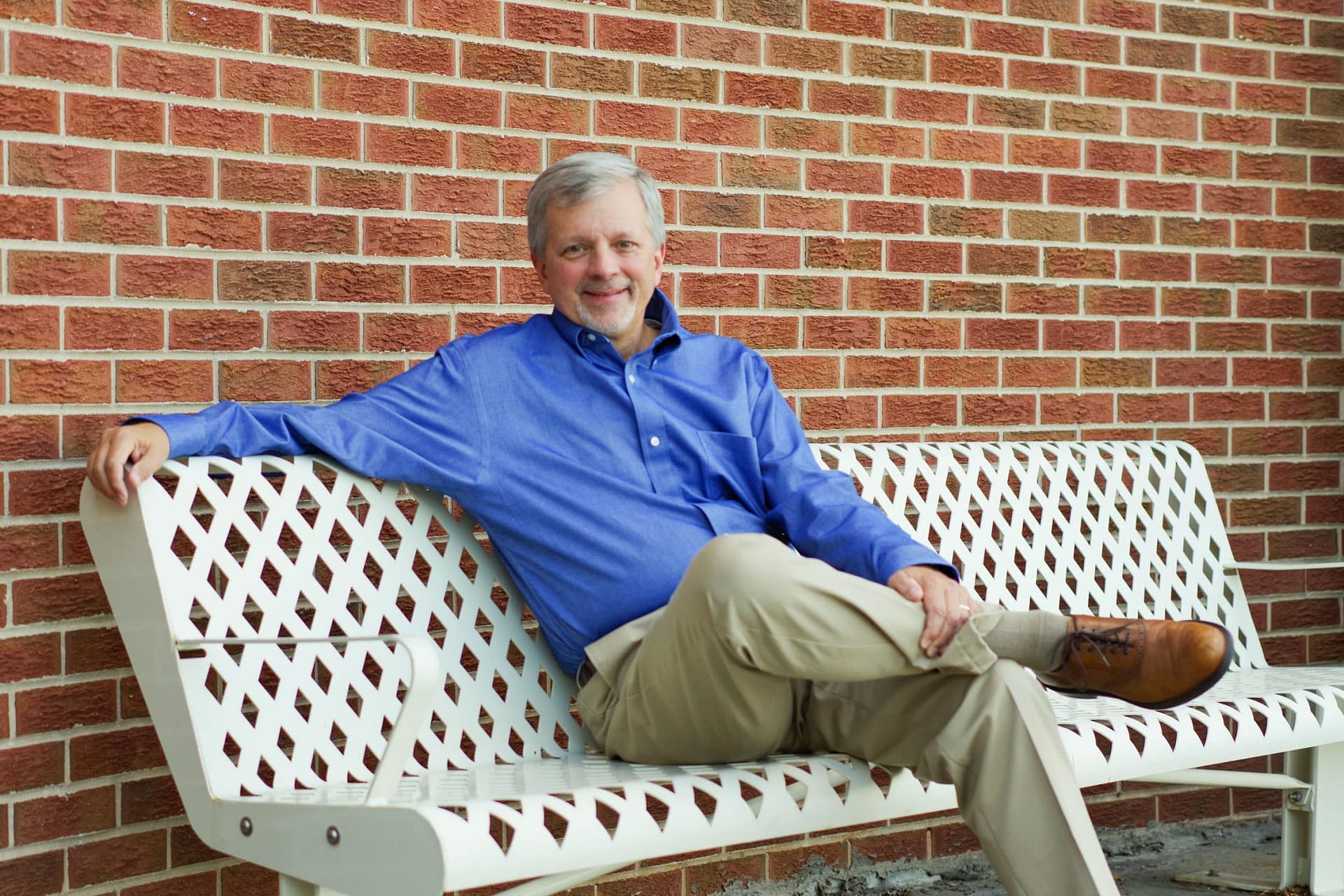 Mark dorn sits on a bench with a brick wall behind him, he is smiling